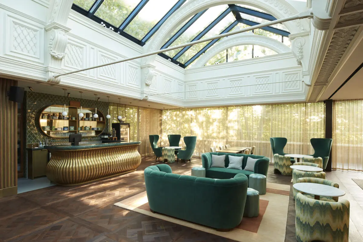 A light filled bar with a glass atrium roof and parquet flooring. The bar is to the left and is gold with a green marble top and the furniture is made up of green velvet sofas and arm chairs and green and white patterned tub chairs.
