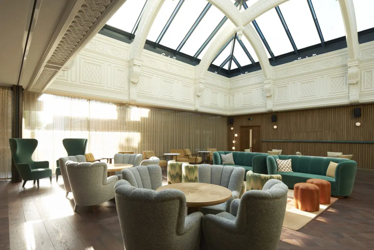 A room with soft chairs in teal, grey and yellow around small wooden round tables and orange footstools