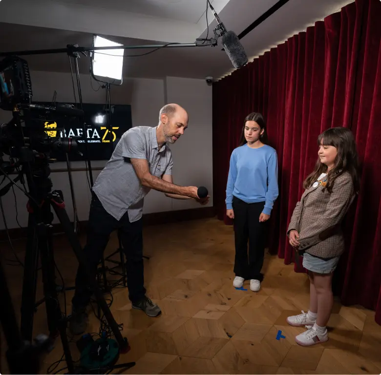 A white man with shaved greying hair and a beard, wearing a short sleeved blue shirt. He si showing two young girls and microphone and they are surrounded by filming equipment.
