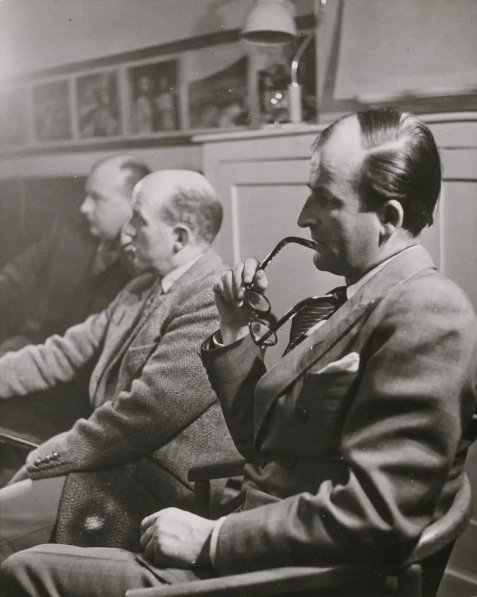Three men wearing suits, the one closest to camera is holding a pair of glasses.