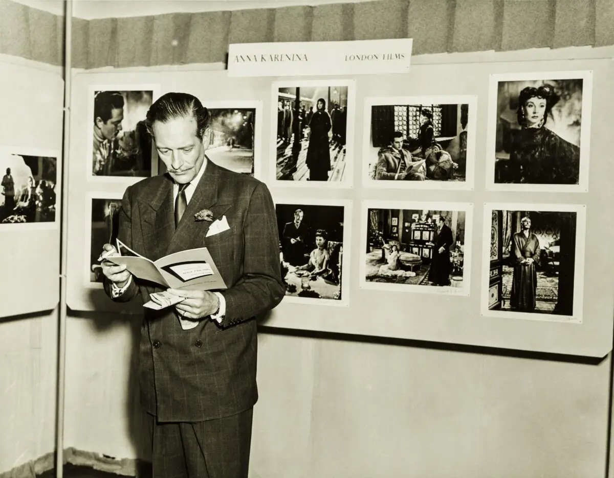 A man with slicked back hair and a thin moustache wearing a suit, reading a booklet and standing in front of a display of screenshots from Anna Karenina.