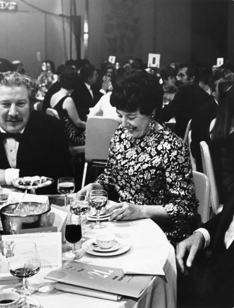A woman with short hair wearing a floral dress is sat at a table and looking down while smiling.