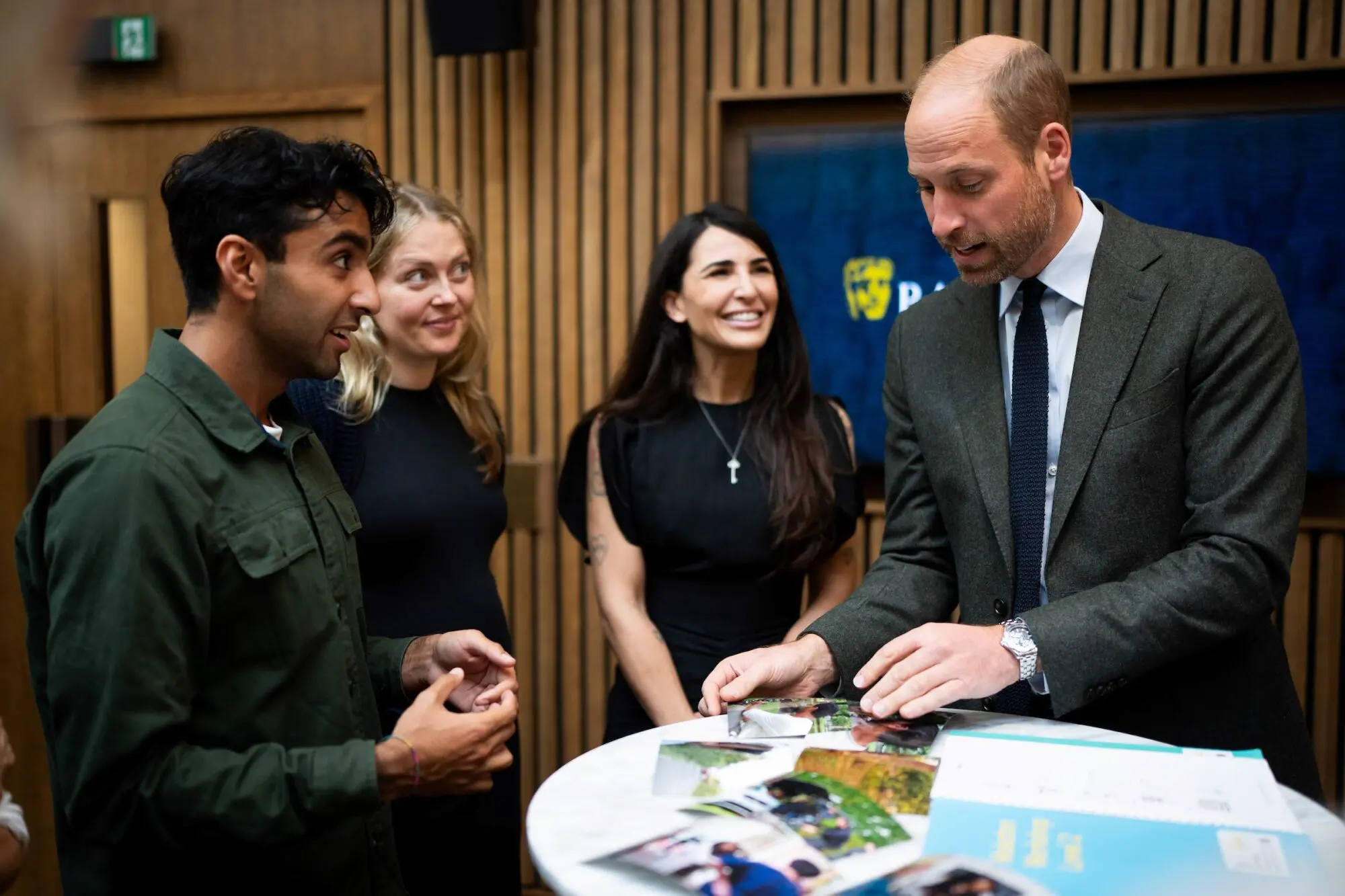 Two men and two women stood around a tall round table looking at brochures. The man on the far right is His Royal Highness, The Prince of Wales.