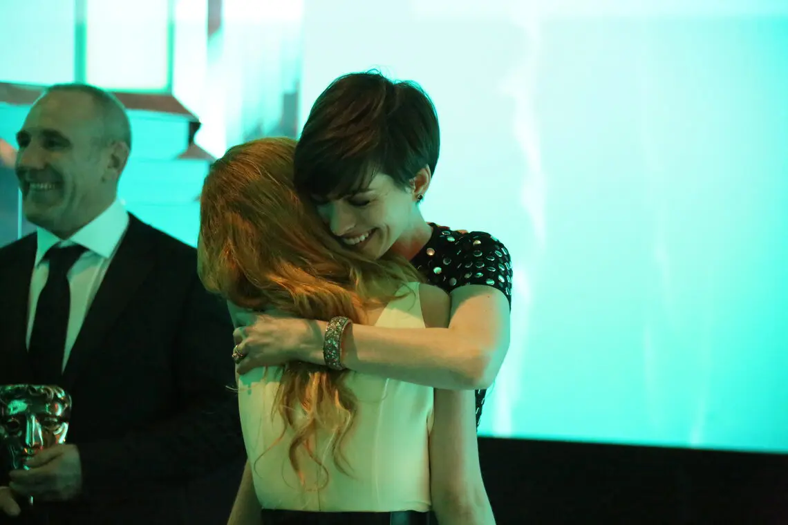 Two women embracing. One has long blonde wavy hair and one has a short dark pixie cut.