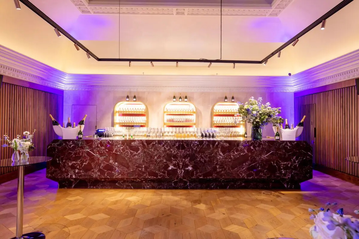 A dark marble bar in front of three backlit alcoves filled with glassware. On top of the bar are several wine and champagne glasses, buckets of champagne and a bouquet of flowers in a glass vase.