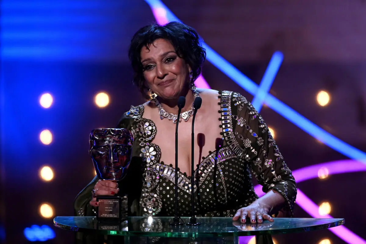 An Indian woman with bobbed black hair and wearing a brown jewelled dress and silver statement collar necklace. She is stood at a lectern holding a BAFTA mask.