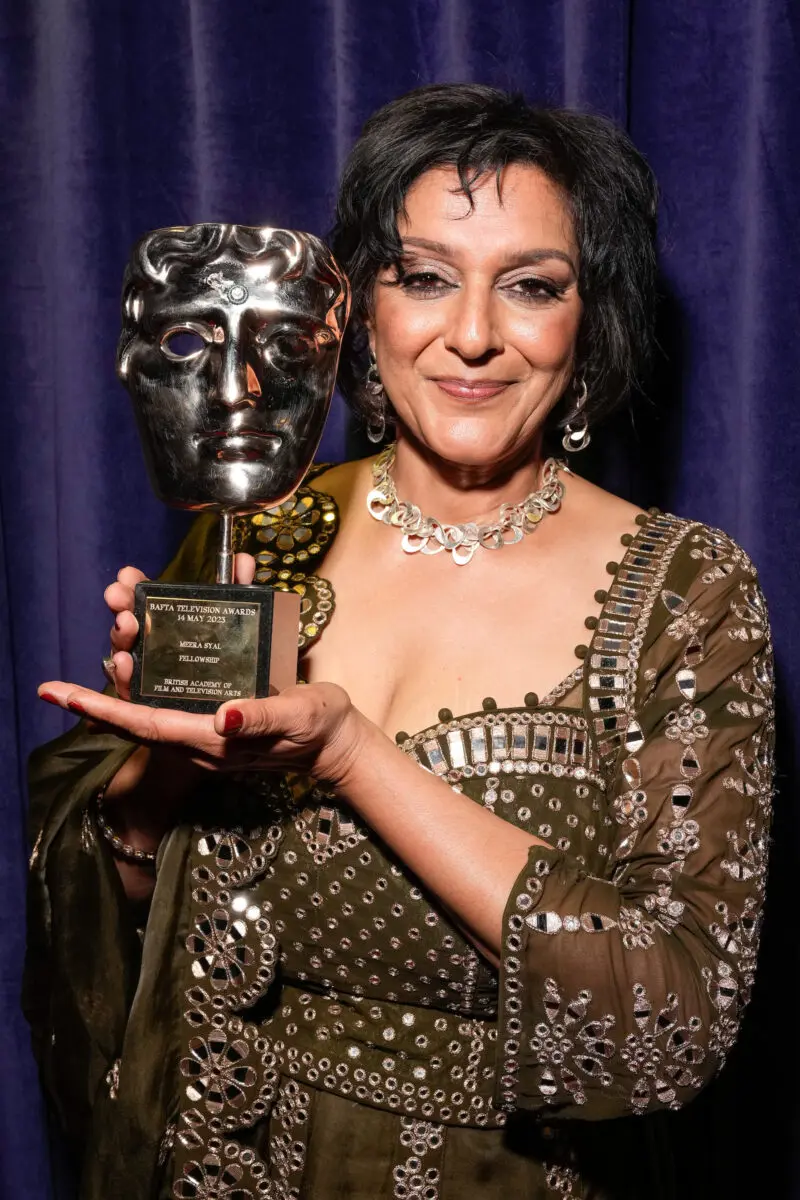 An Indian woman with bobbed black hair and wearing a brown jewelled dress and silver statement collar necklace. She is stood in front of a blue curtain holding a BAFTA mask.