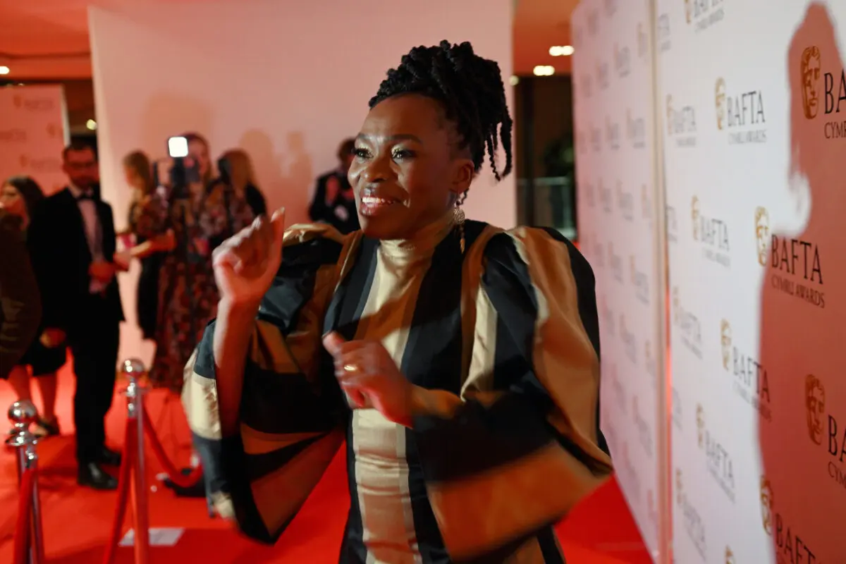 A black woman with braids in a high bun dancing on the red carpet. She wears a gold and black striped dress.