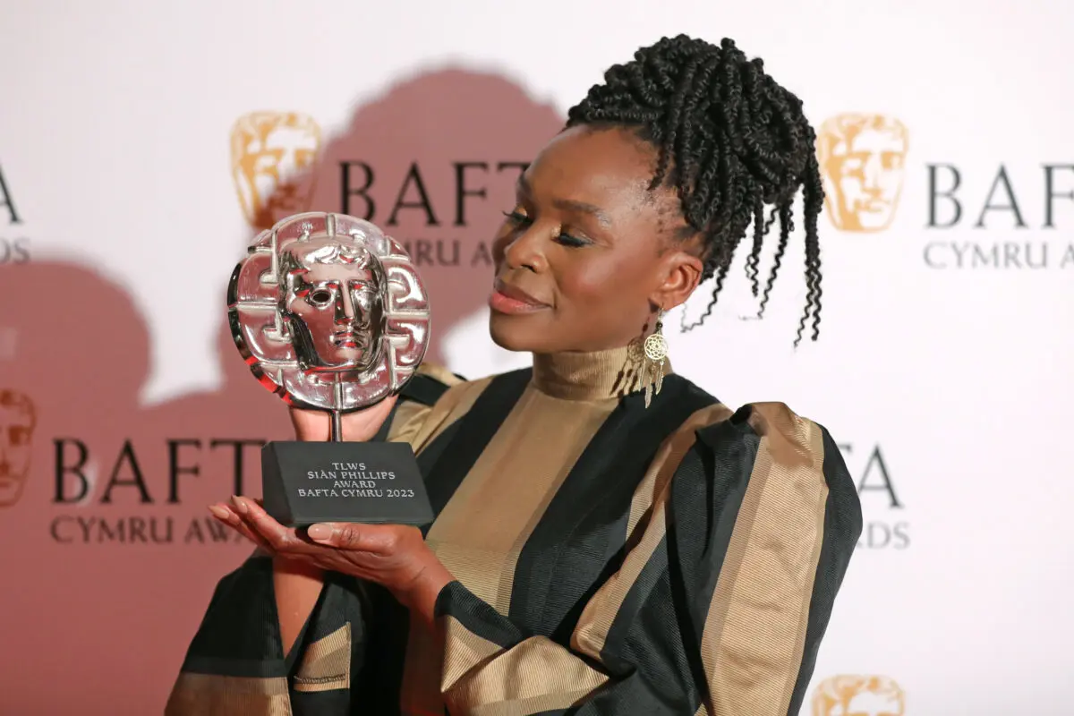A black woman with braids in a high bun poses with a BAFTA award. She is wearing a gold and black striped dress.