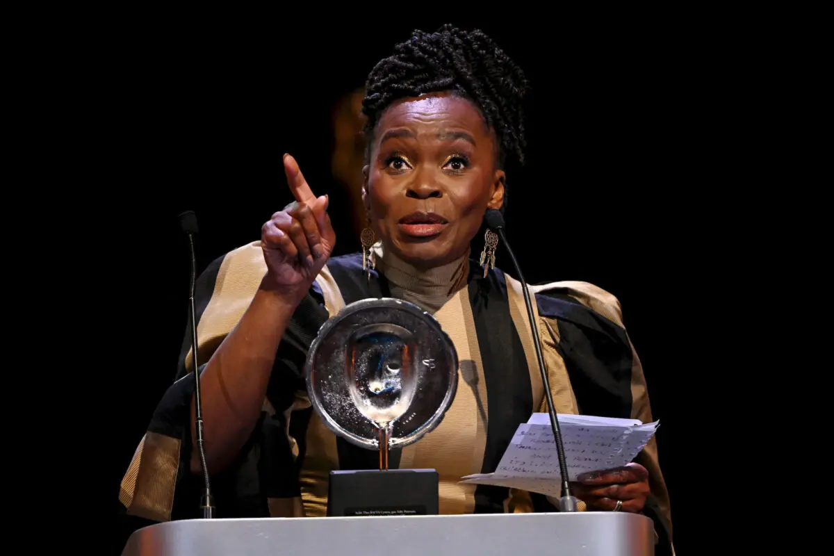 A black woman with braids in a high bun collects an award on stage. She is holding some white paper and pointing at the crowd. She is wearing a gold and black striped dress.