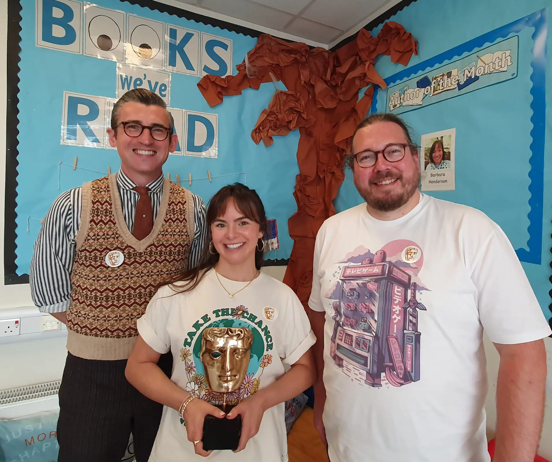 A woman with dark hair holding a BAFTA award and stood between two men, one in a sweater vest, in a classroom.