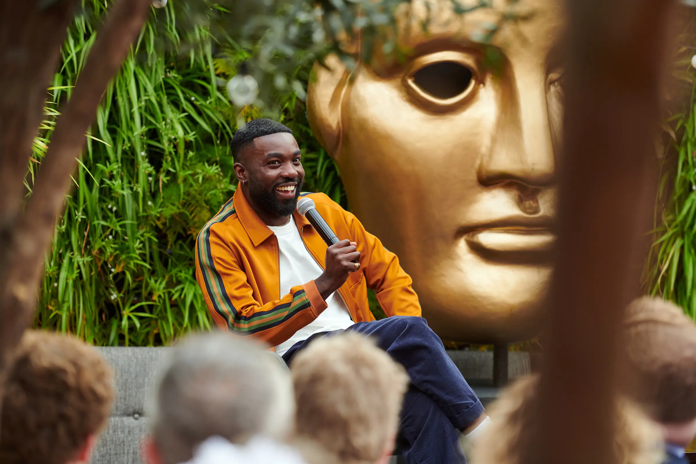 A black man in a white t-shirt and orange tracksuit jacket speaking into a microphone.