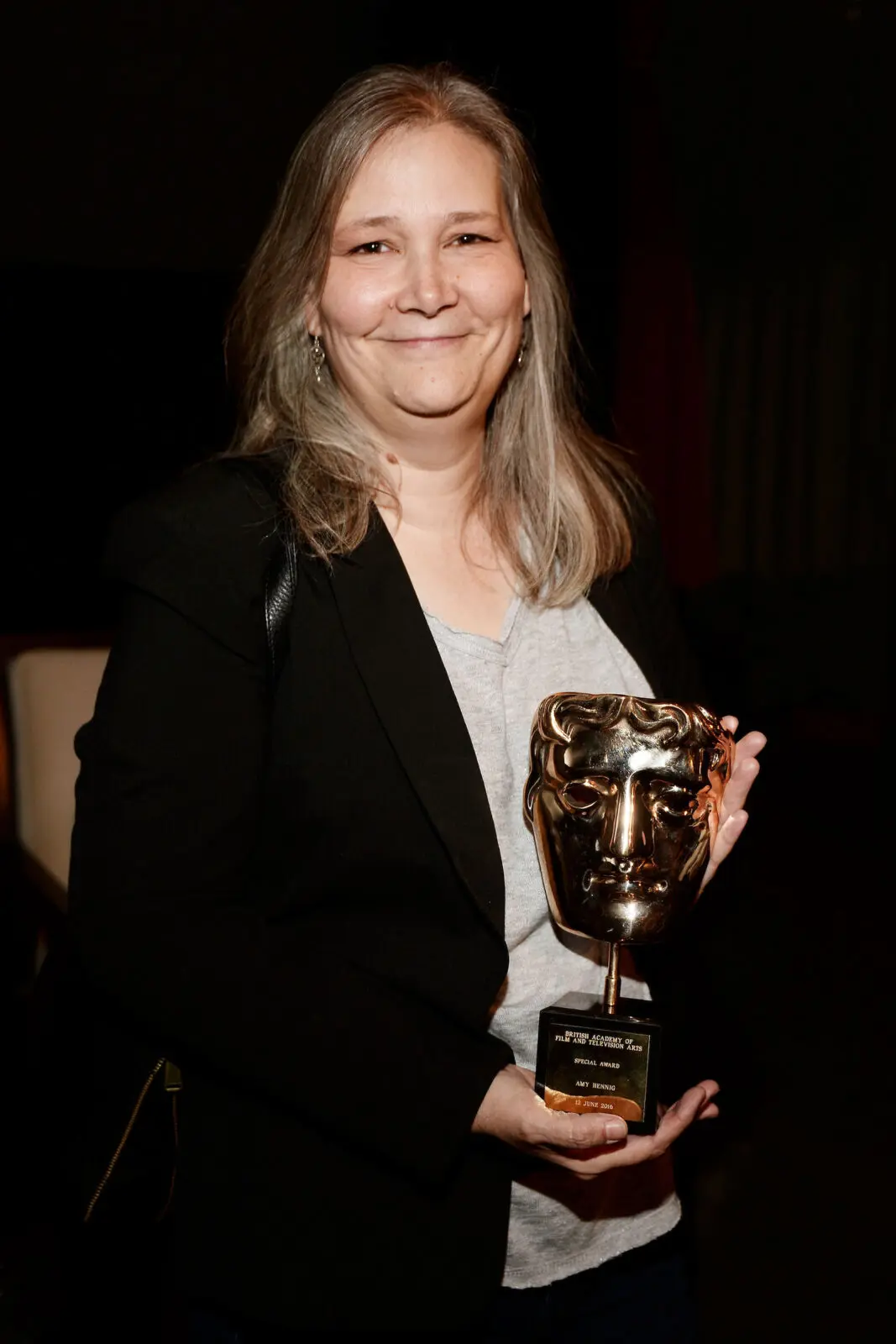 A white woman with shoulder length greying hair. She is wearing a grey t-shirt and black jacket and is holding a BAFTA award.