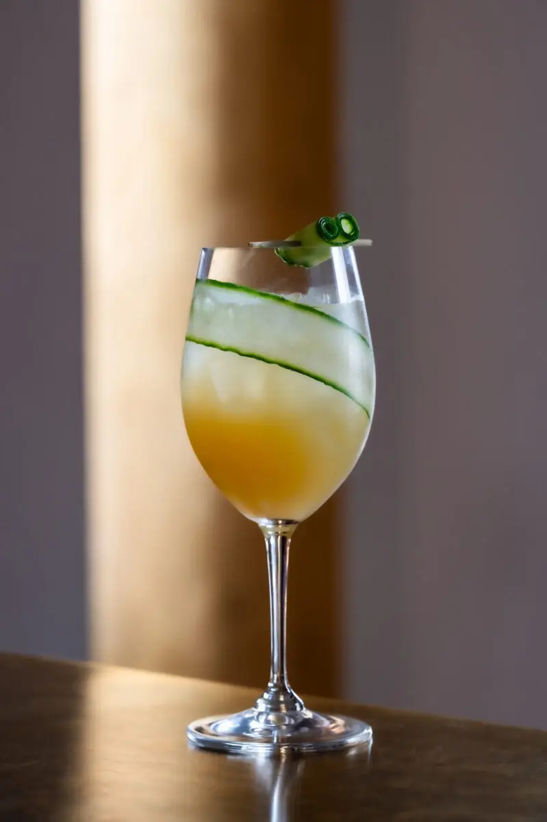 A tall glass with orange liquid in and a curl of cucumber.