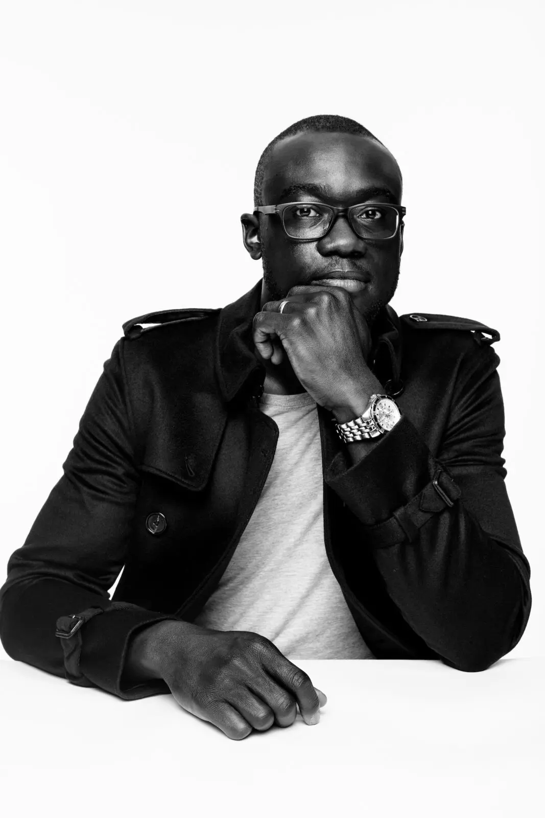 A black man with short cropped hair and thick rimmed glasses. He is wearing a grey t-shirt and black trench coat.