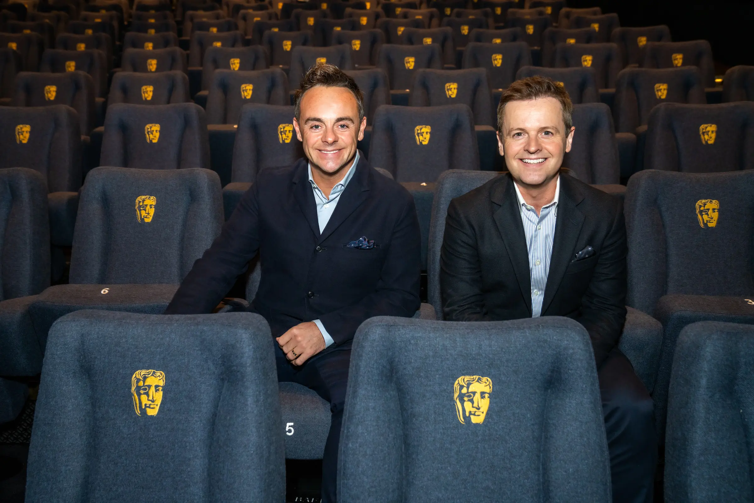Two white men with short dark hair wearing blue shirts and black suit jackets. They are sat in blue cinema seats with the BAFTA logo on.