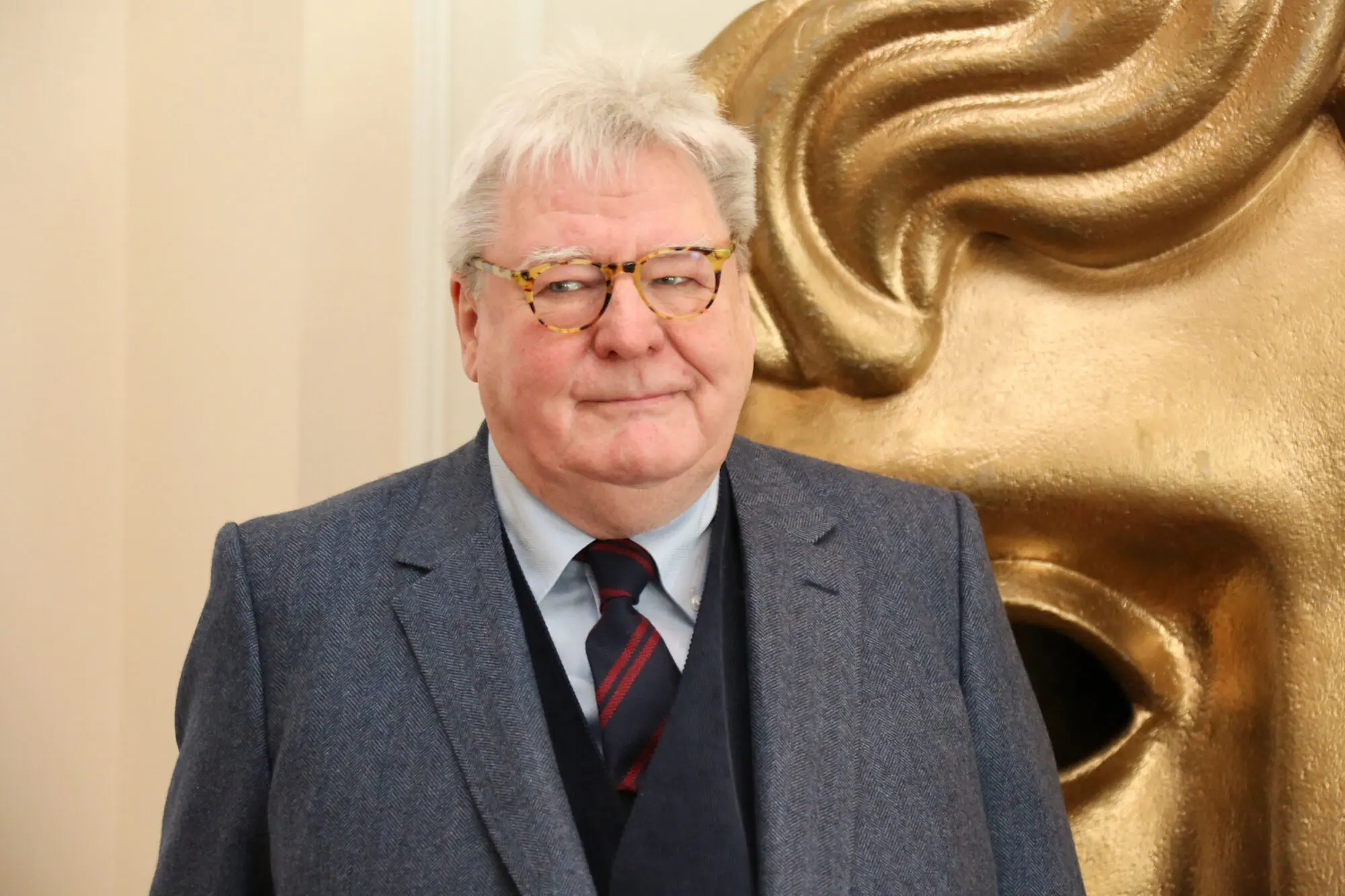 A white man with white hair wearing tortoise rimmed glasses, a pale blue shirt, black waistcoat, tweed blazer and blue and red striped tie.