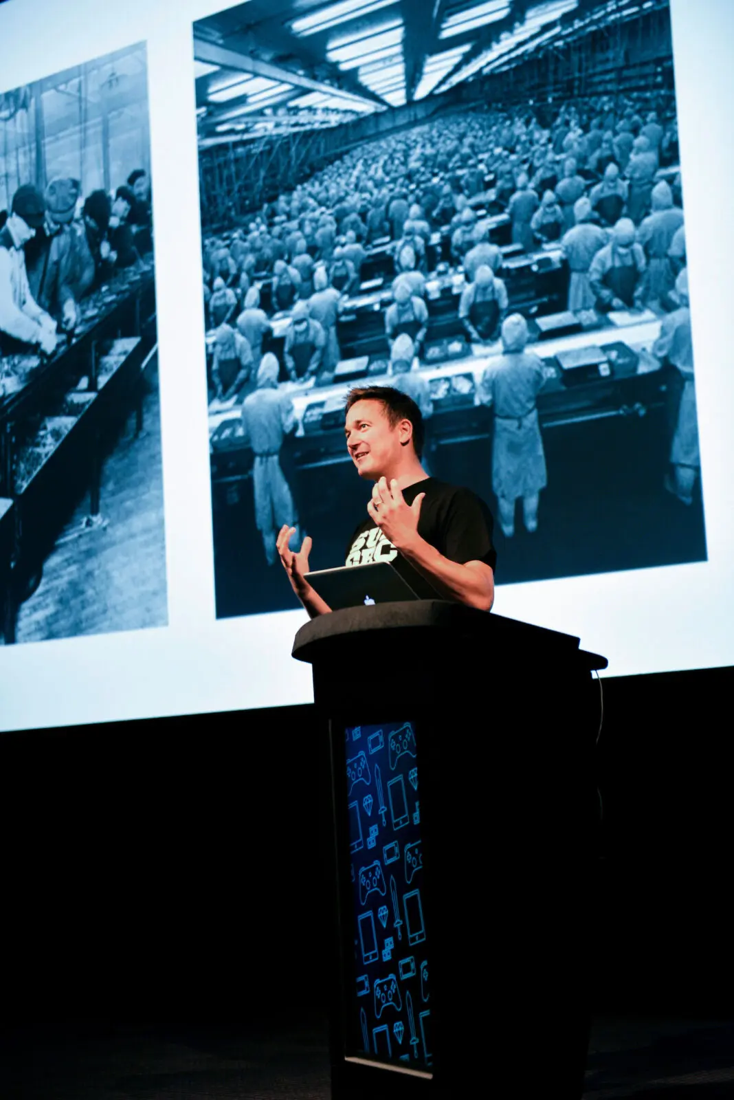 A white man with dark hair, wearing a black t-shirt with white text on, giving a speech at a lectern.