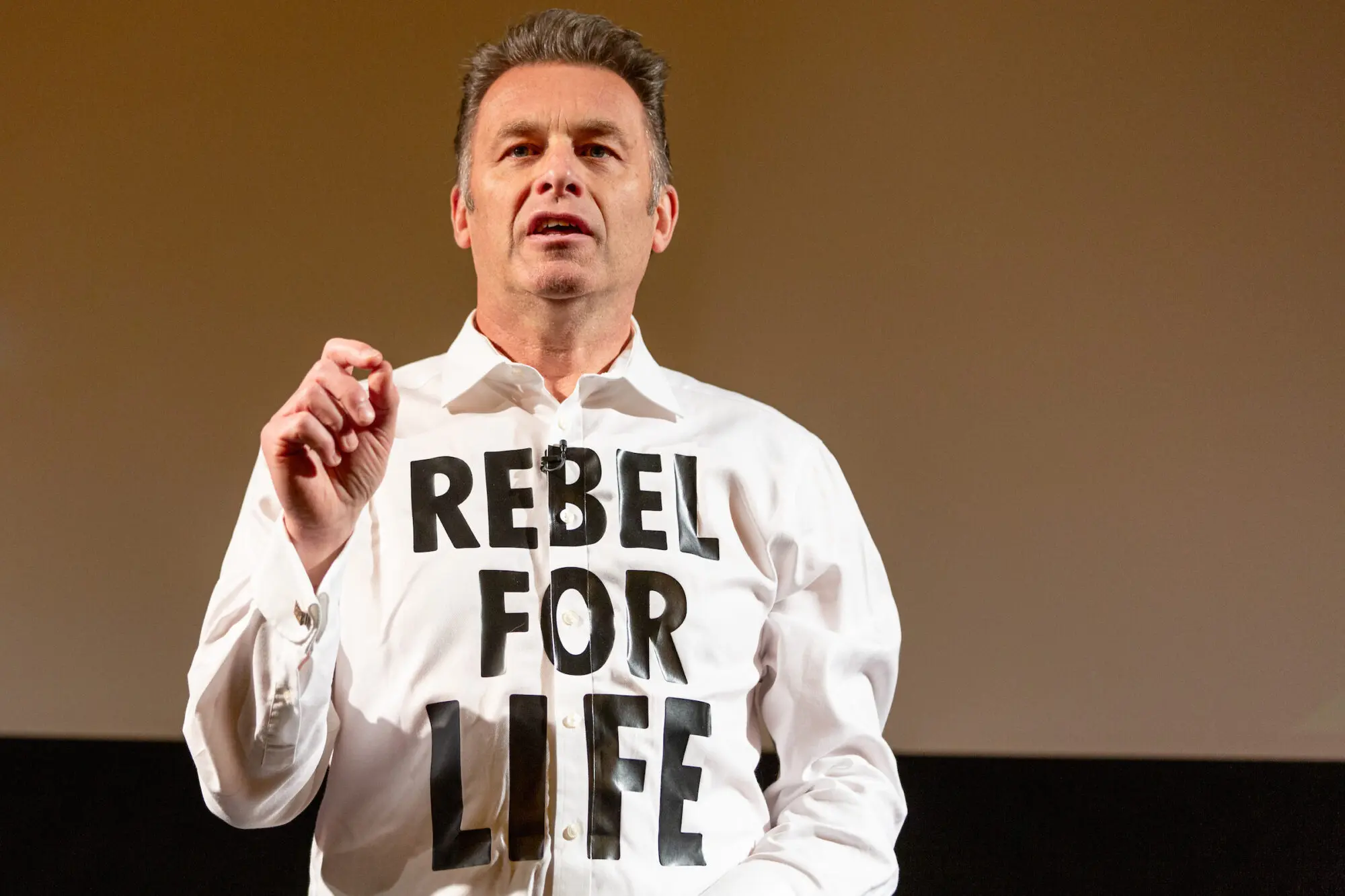 A white man with thick grey hair wearing a white shirt with 'Rebel for Life' written on it.