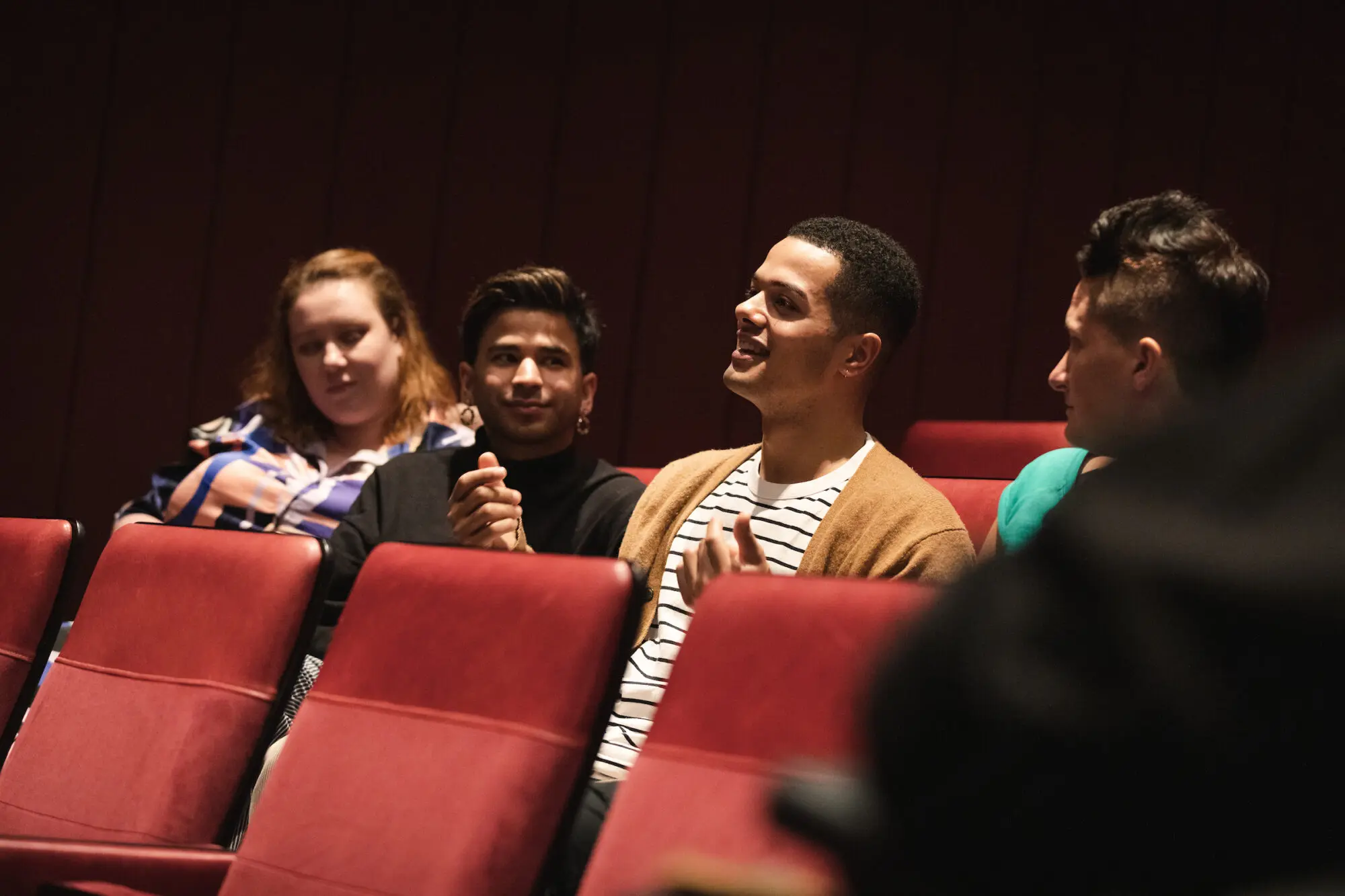 A young man wearing a striped top and tan cardigan sat in a red cinema seat talking.