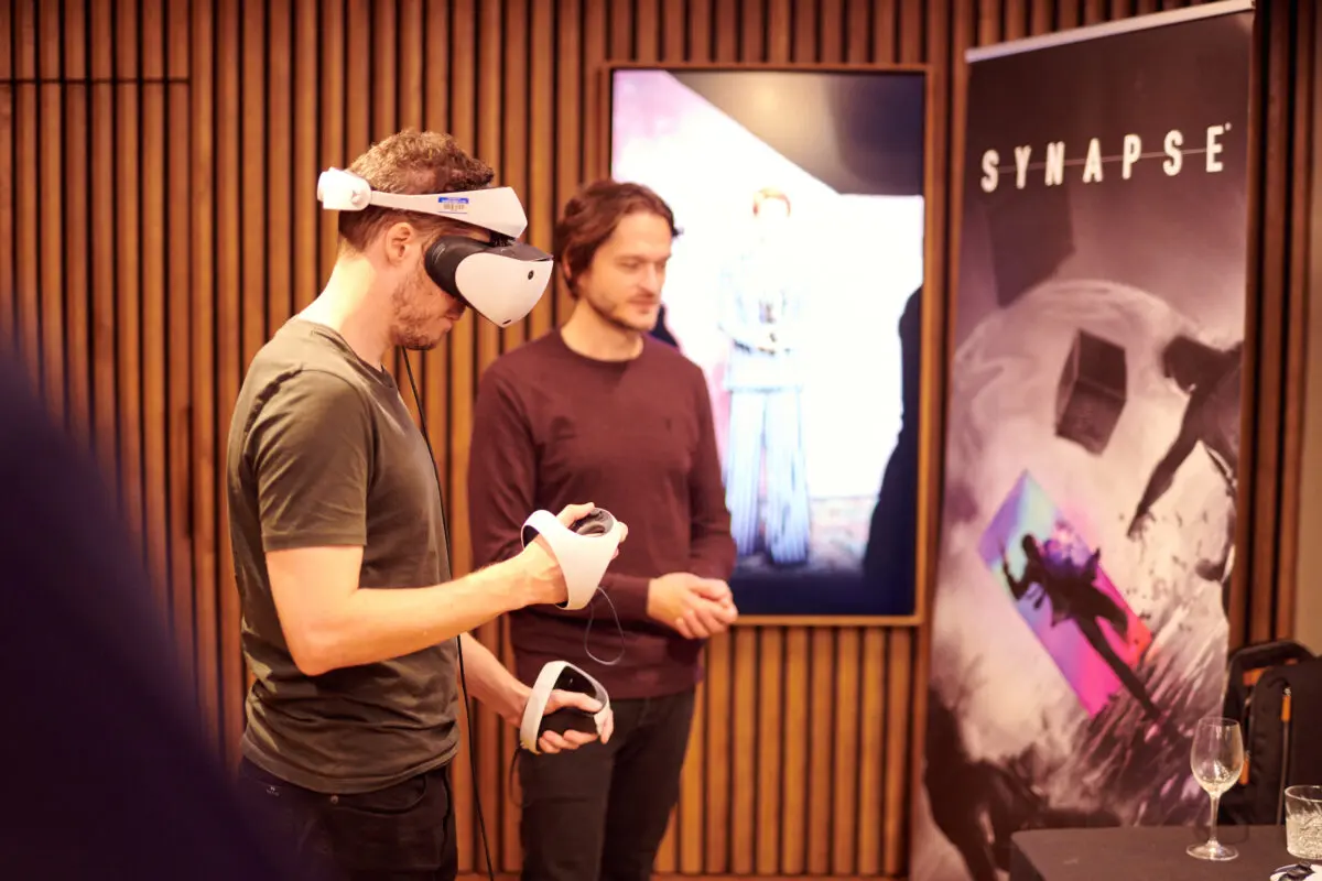 Two men, one with a VR headset on playing a game.