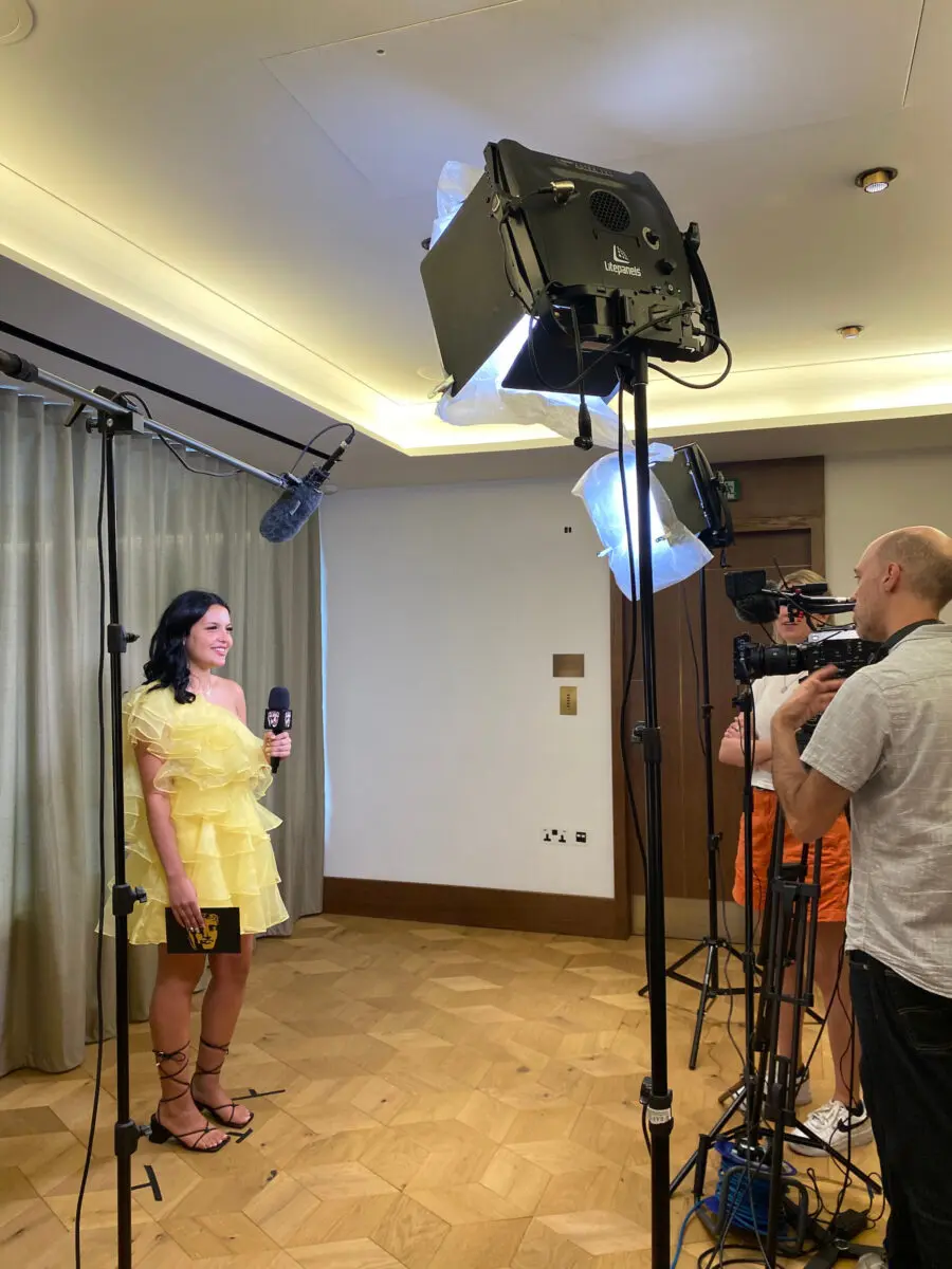 A young woman with dark shoulder-length hair and a yellow dress giving an interview to camera. A large spotlight is shining on her and a cameraman is filming.
