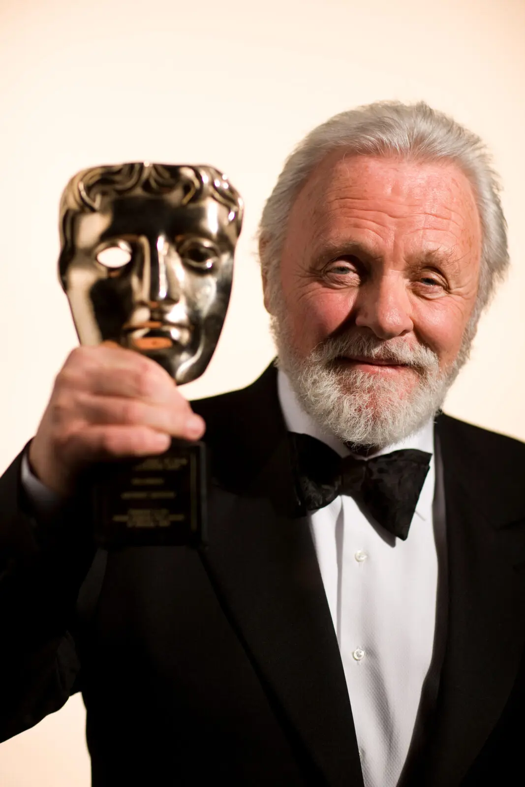 Sir Anthony Hopkins. An older white man with thick white hair and a beard. He is wearing a black tuxedo and bow tie and is holding a BAFTA mask.