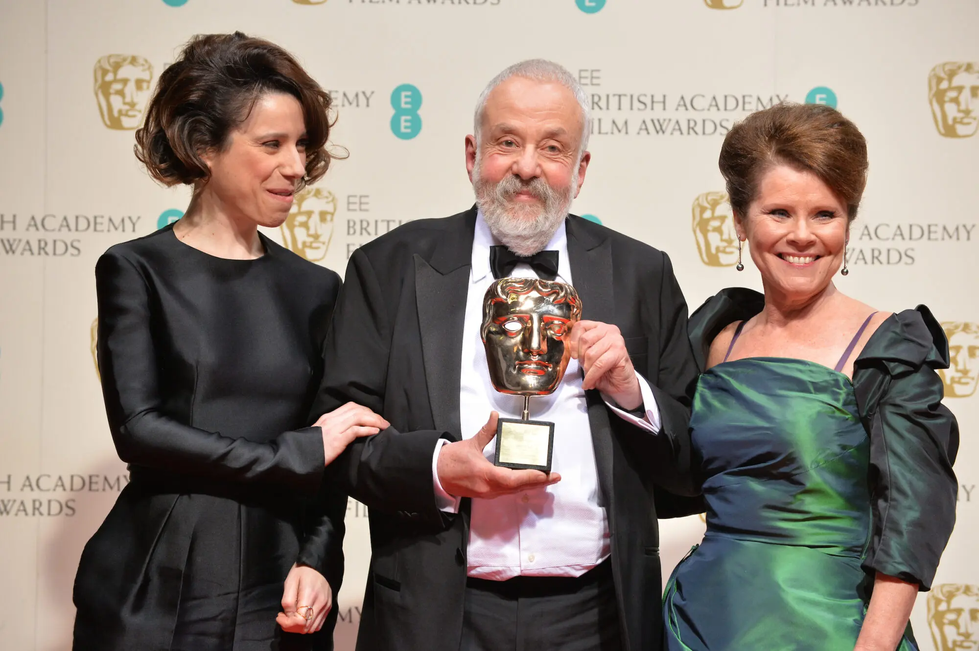 A white man with white hair and beard wearing a tuxedo and bow tie and holding a BAFTA mask. On the left is a woman with bobbed dark hair and wearing a long sleeved black dress and on the right is a woman with brown hair tied up and wearing an iridescent green dress with puffed sleeves.