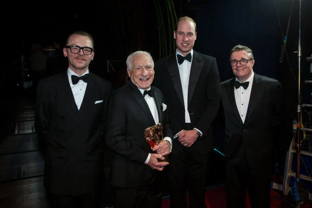 Four white men in tuxedos. From left to right; a man with short dark hair wearing thick rimmed black glasses, an older man with white hair holding a BAFTA mask, a tall man with dark blond hair and a man with thick dark hair with white streaks in the side and thick rimmed dark glasses.