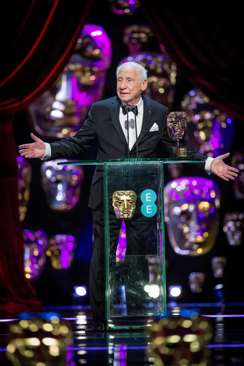An older white man with white hair stood at a lectern on a stage with a BAFTA mask.