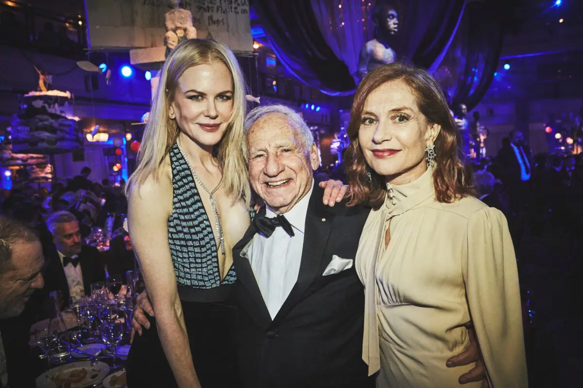 An old white man with white hair and wearing a black tuxedo and bow tie stood with his arms around two women. The woman on the left has long blonde hair and is wearing a low cut black and silver dress with a long diamond necklace. On the other side of him is a woman with bobbed brown hair and red lipstick wearing a beige pussy bow top and sparkly star earrings.