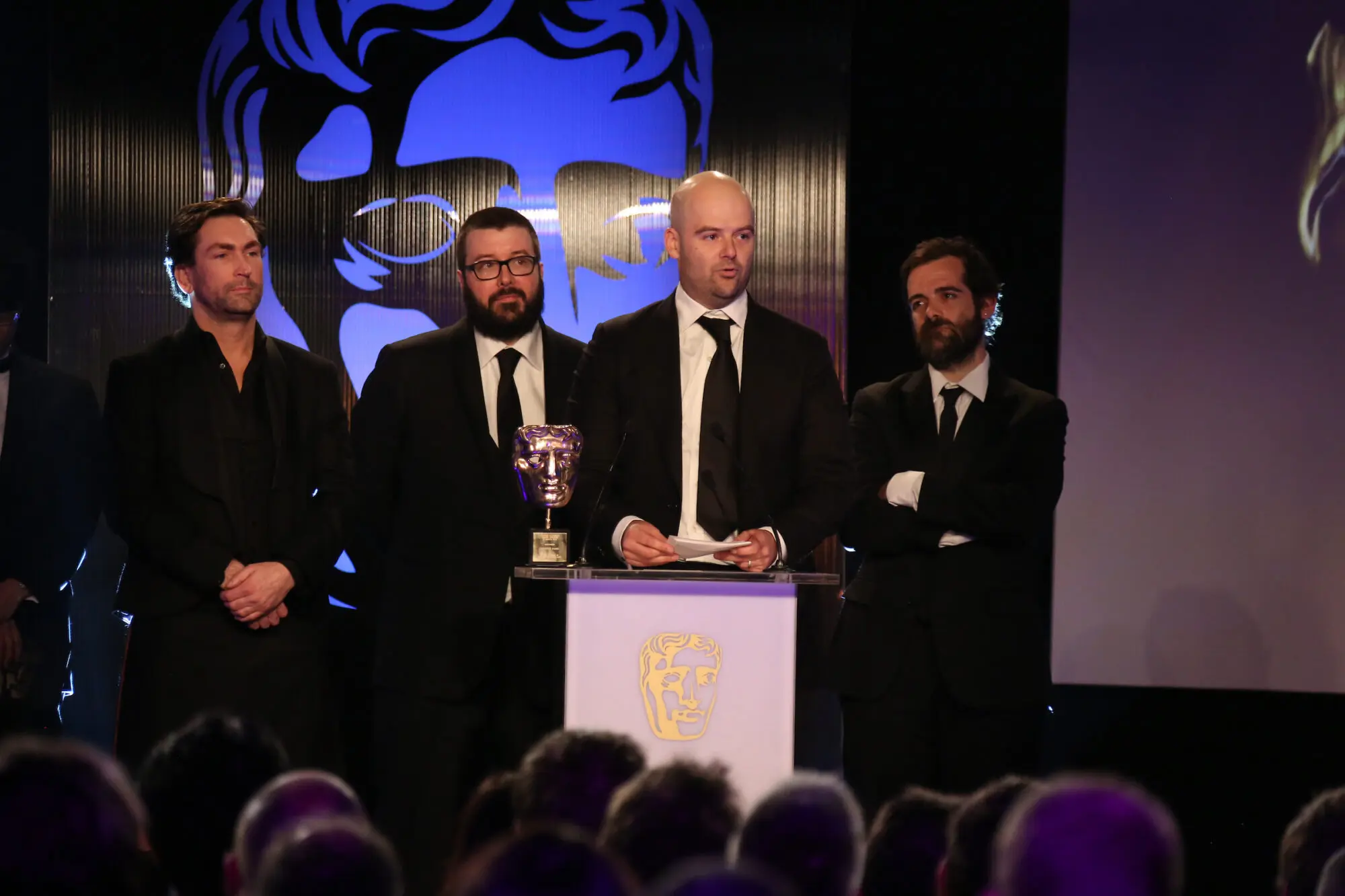 Four white men stood at a BAFTA branded lectern which has a BAFTA mask on it. They are all wearing black suits.