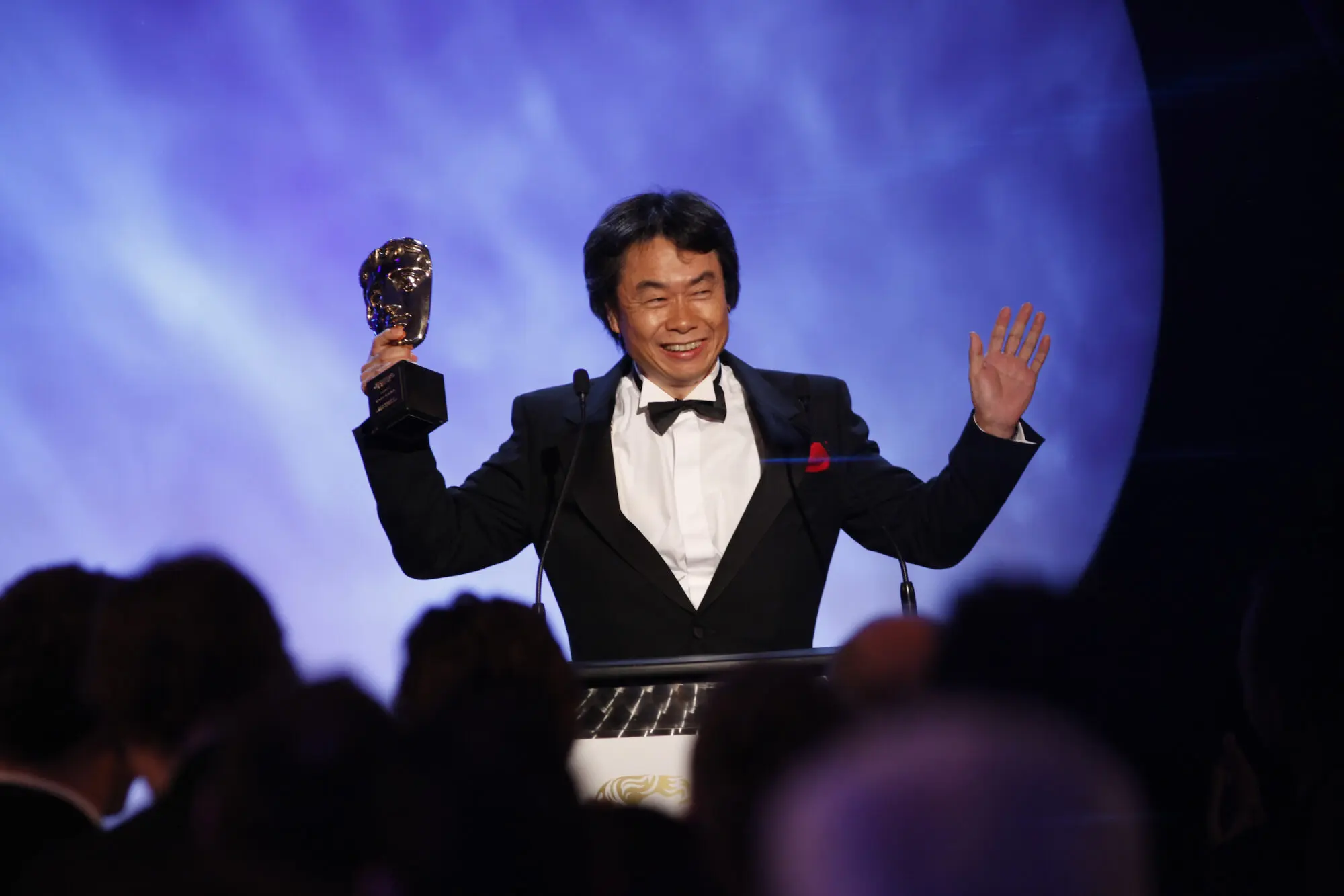 An Asian man with thick black hair wearing a black suit jacket, black bow tie and red pocket square. He is holding a BAFTA mask.
