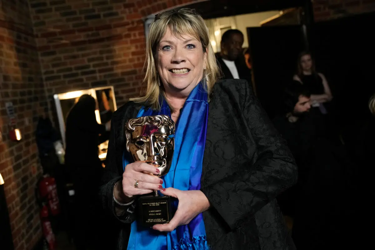 A white woman with a blonde bob and fringe. She is wearing a black jacket with silky blue scarf and glasses on her head. She is posing backstage with a BAFTA mask.