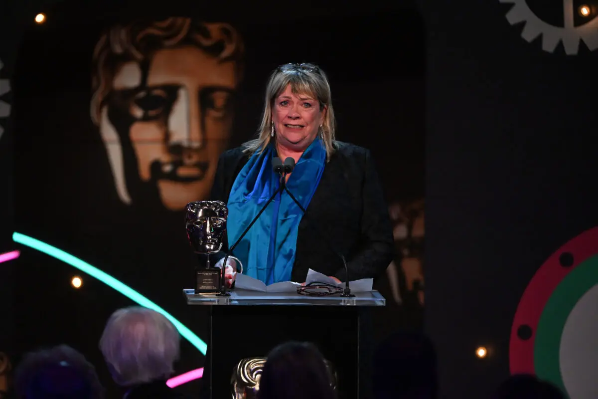 A white woman with a blonde bob and fringe. She is wearing a black jacket with silky blue scarf and glasses on her head. She is standing at a lectern with a BAFTA mask on.