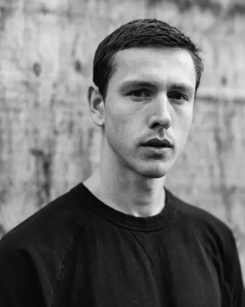 A black and white photo of a young white man with dark hair staring straight at the camera. He is wearing a plain black t-shirt.