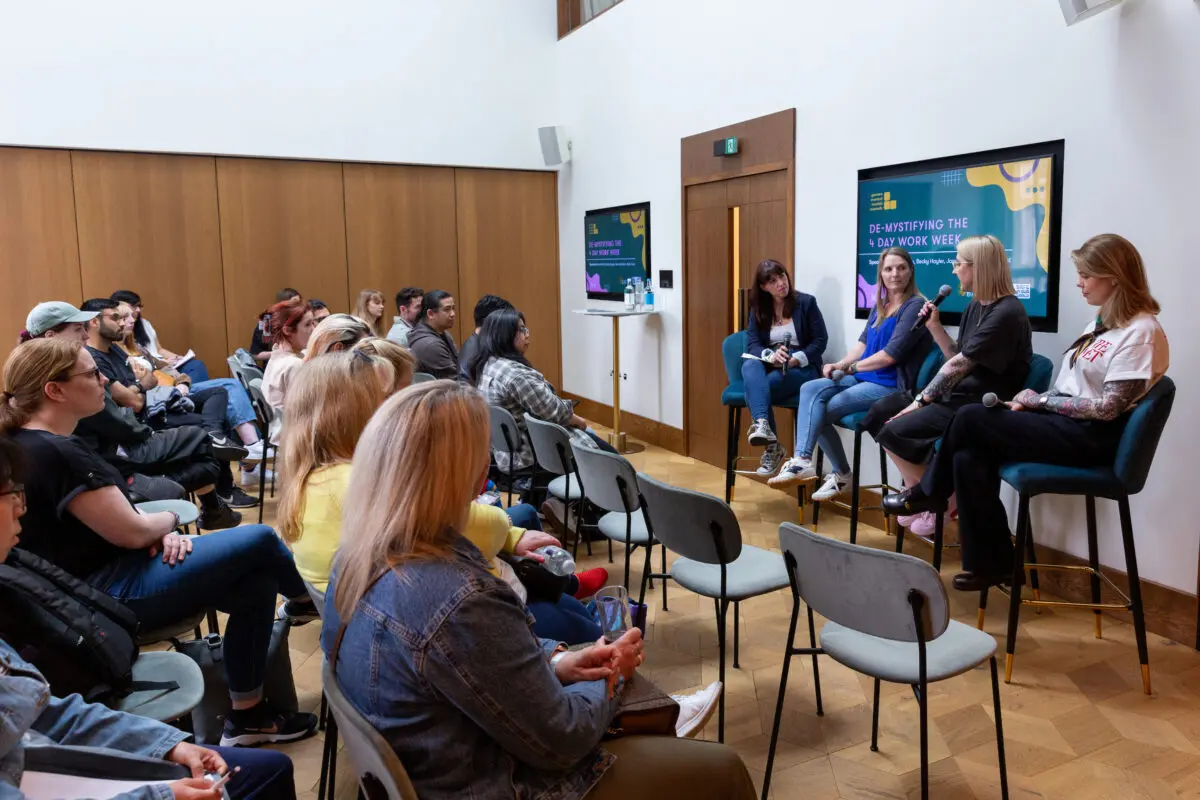 People sat watching a panel of three people sat on high chairs and leading a discussion.
