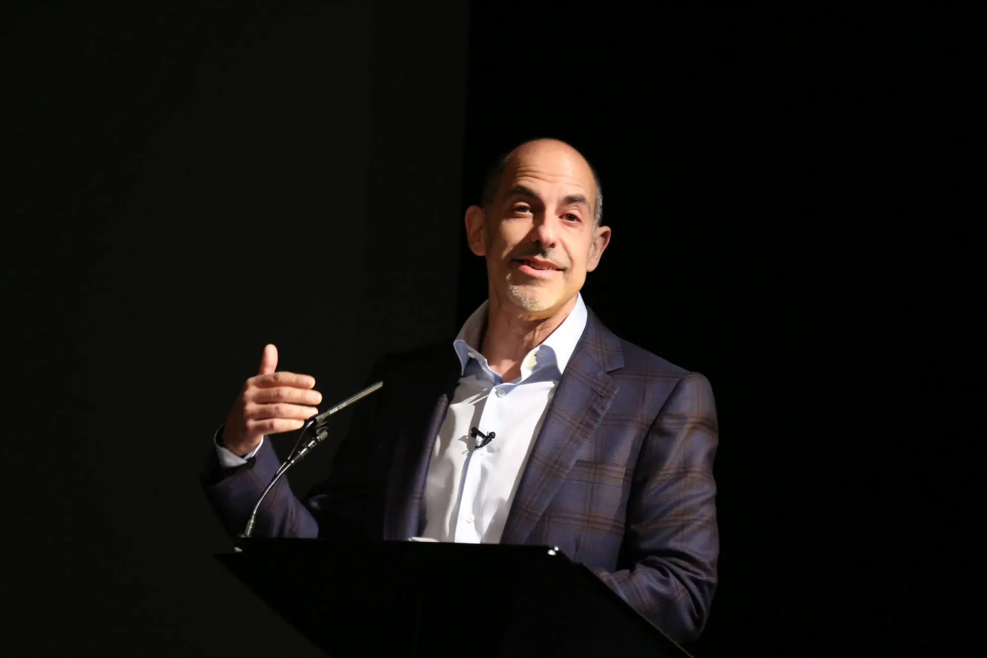 A white man with grey hair on the sides. He is wearing a dark blue plaid suit jacket.