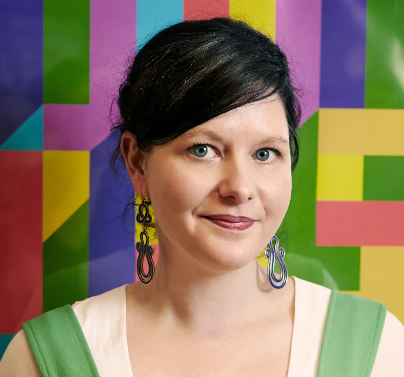 A white woman with black hair tied back and swept across her forehead. She is wearing a white top with green straps and large statement earrings. Behind her is a colourful patterned wall.