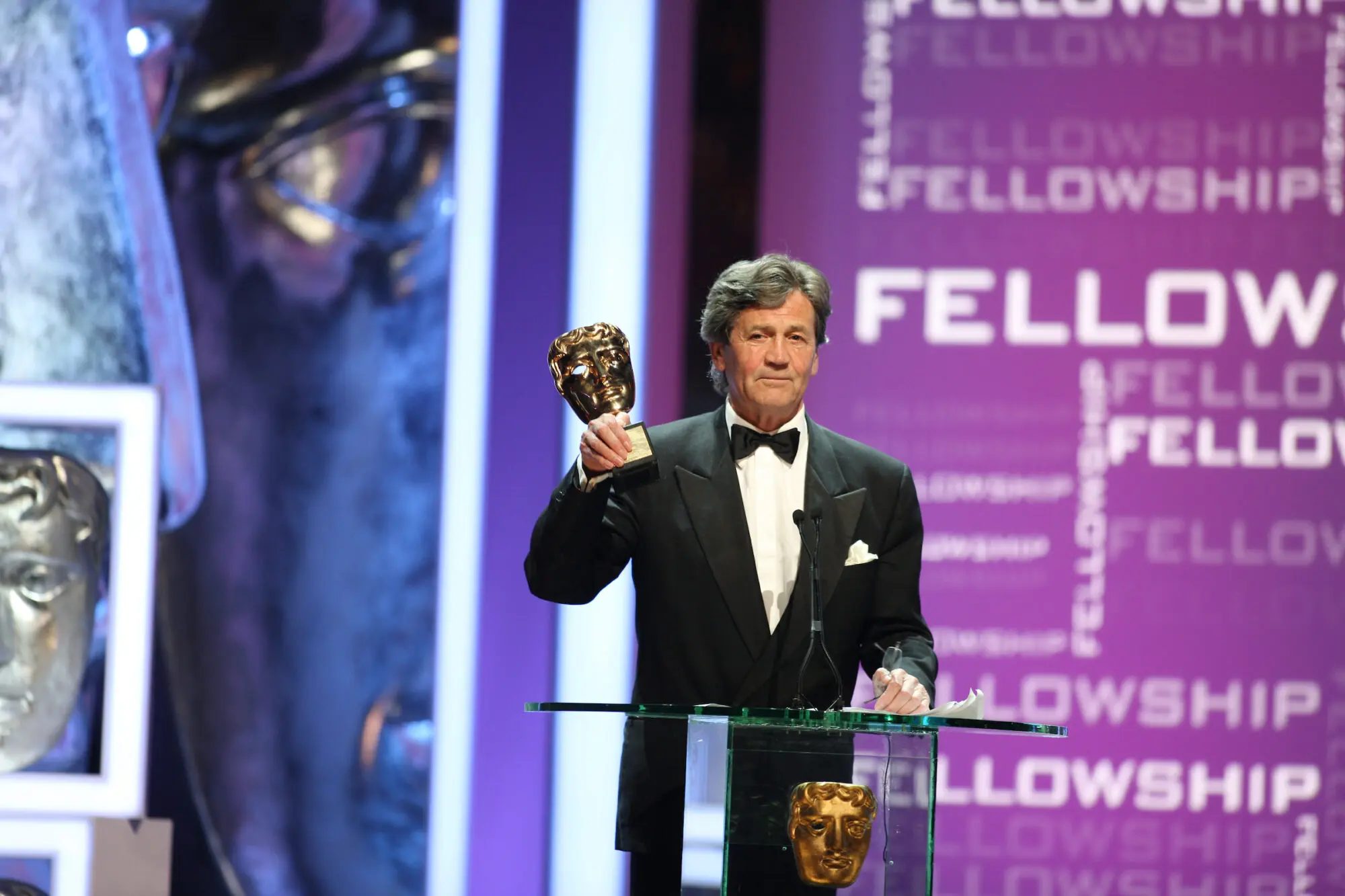 A white man with thick dark grey hair. He is wearing a black tuxedo with a bow tie and white pocket square. He is stood on stage holding a BAFTA award.