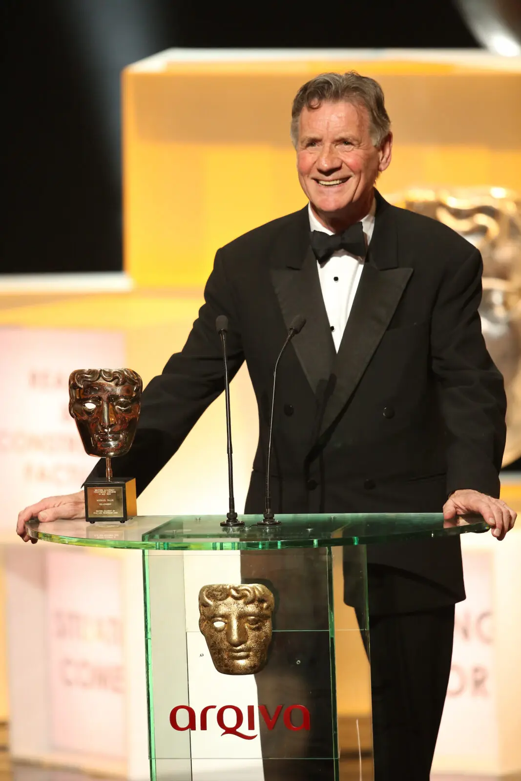 A white man with dark grey hair with a curl at the front. He is wearing a black tuxedo and bow tie and is stood on a stage with a BAFTA award.