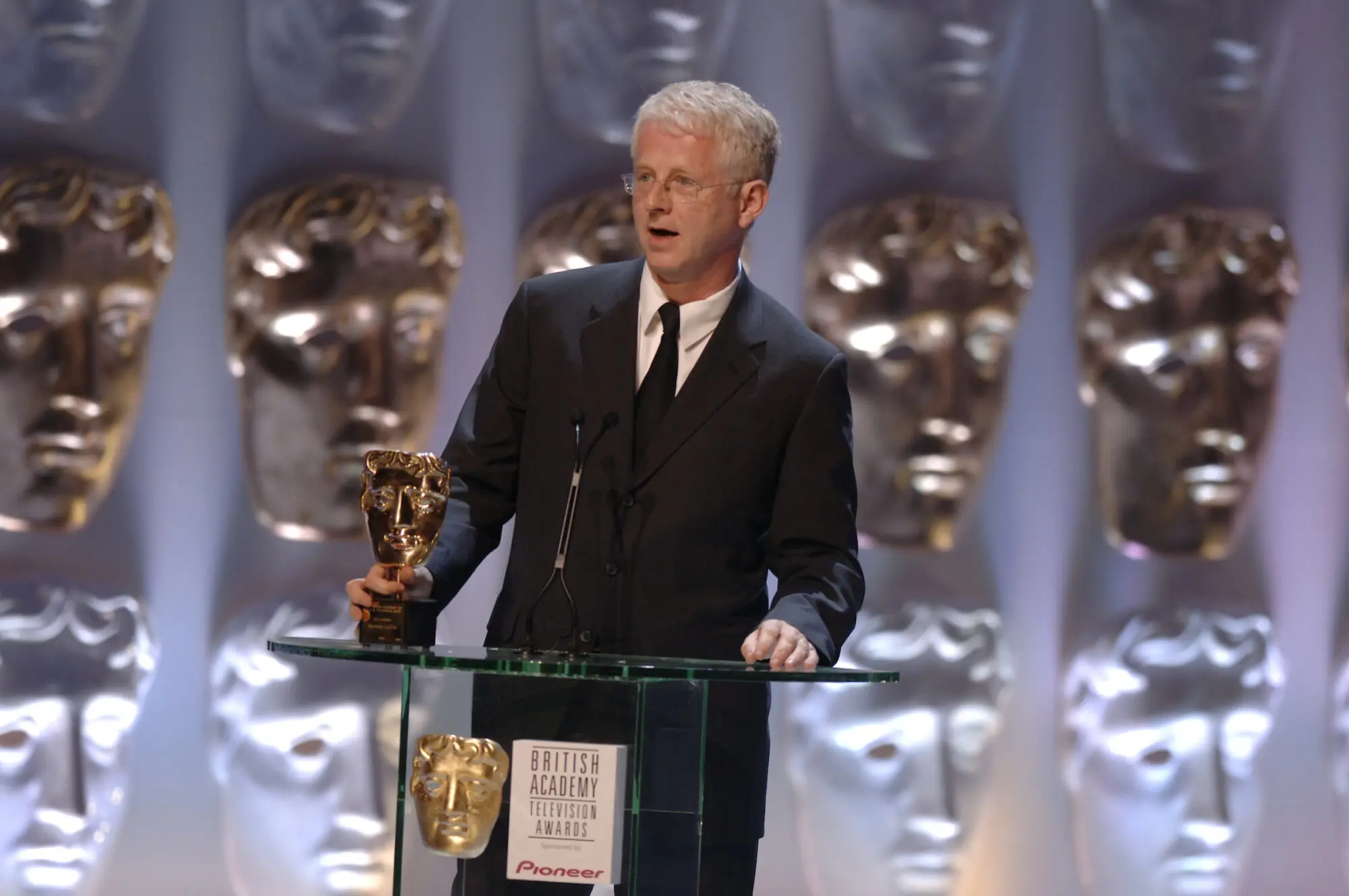 A white man with short white hair and small frameless glasses. he is wearing a black suit and tie and holding a BAFTA mask.