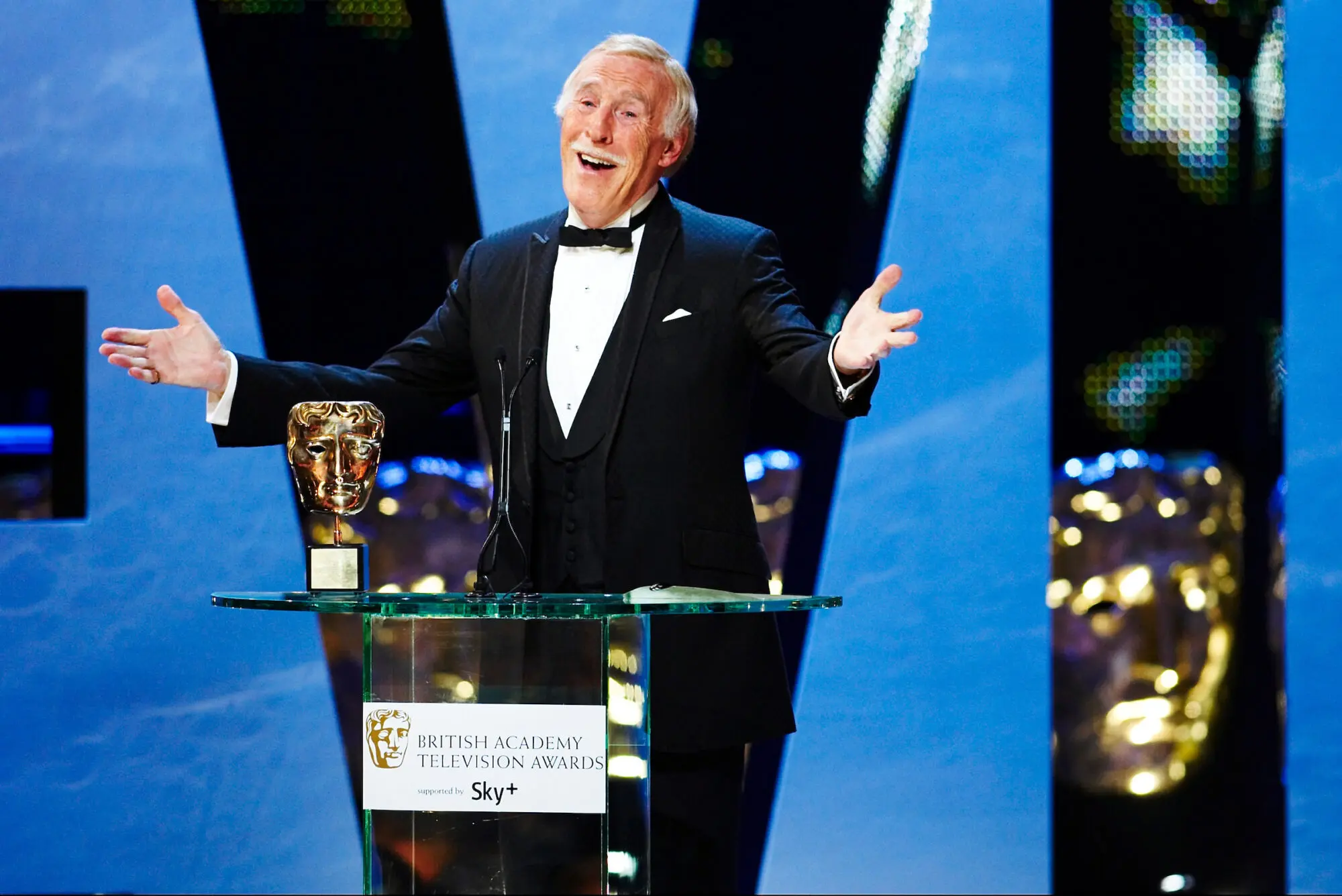 An old white haired man with a white moustache in a black tuxedo. His arms are stretched out as he stands behind a glass podium with a BAFTA placed on it