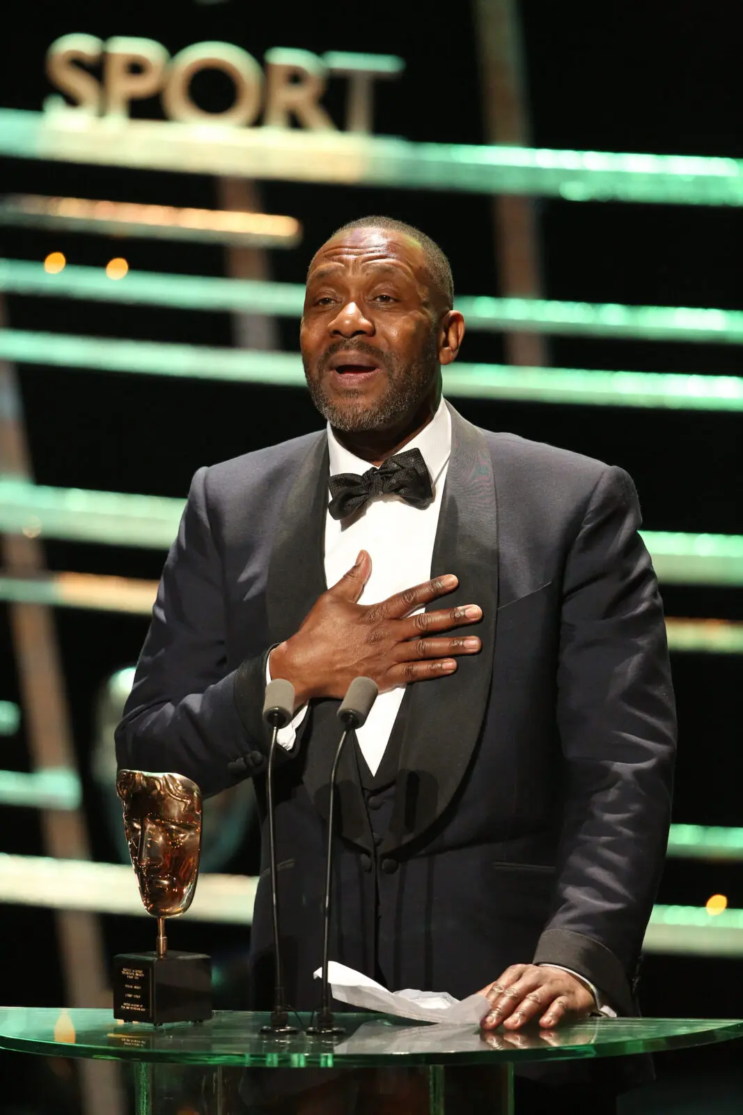 A black man with short shaved hair and dark stubble. He is wearing a tuxedo and black bow tie and is giving a speech with a BAFTA mask in front of him.