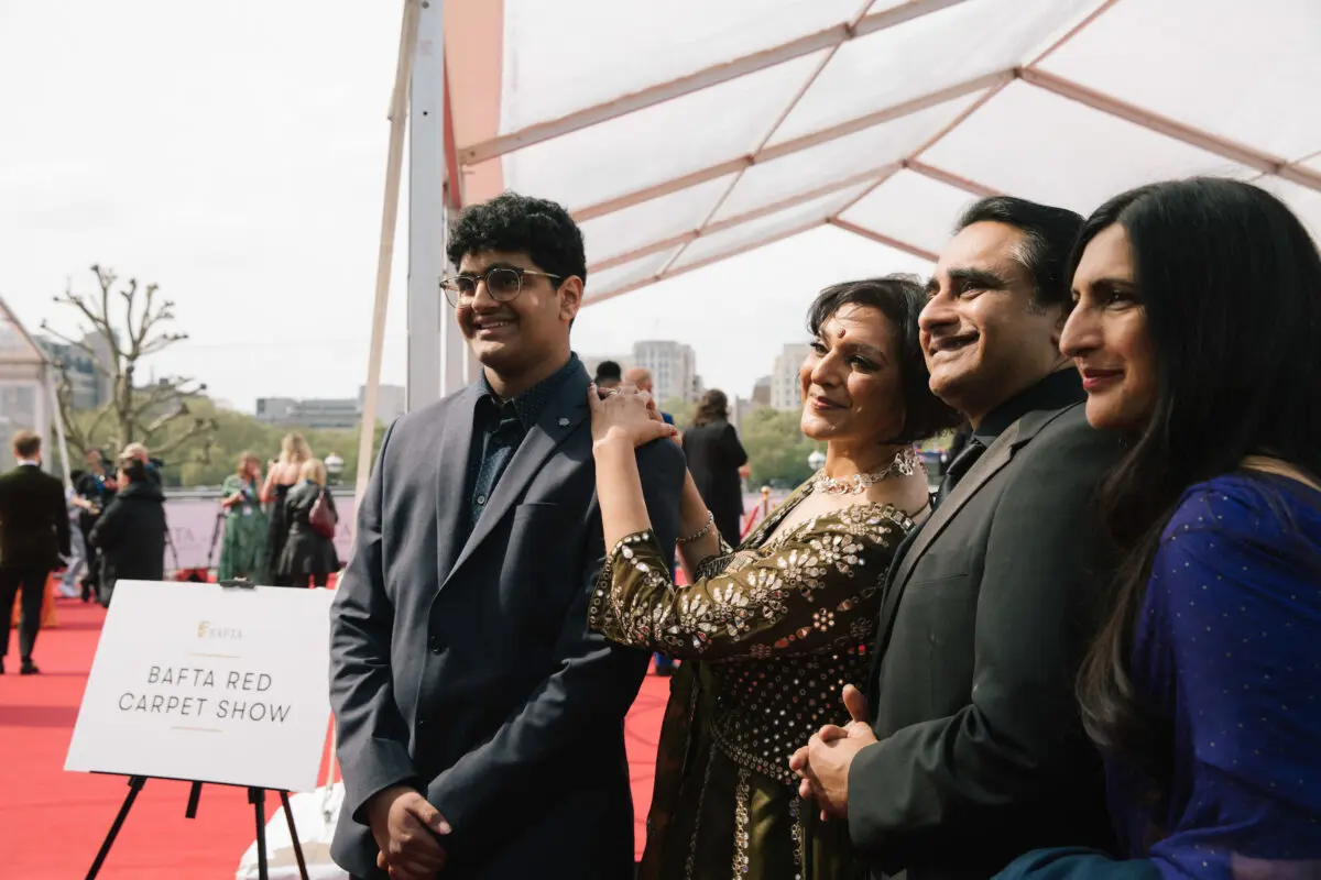 Four Indian people stood posing on the red carpet together. From left to right there is a young man with dark hair and glasses in a grey suit, a woman with bobbed black hair and a jewelled dress, a man with thick dark hair and a dark suit and a woman with long dark hair and wearing a blue dress.