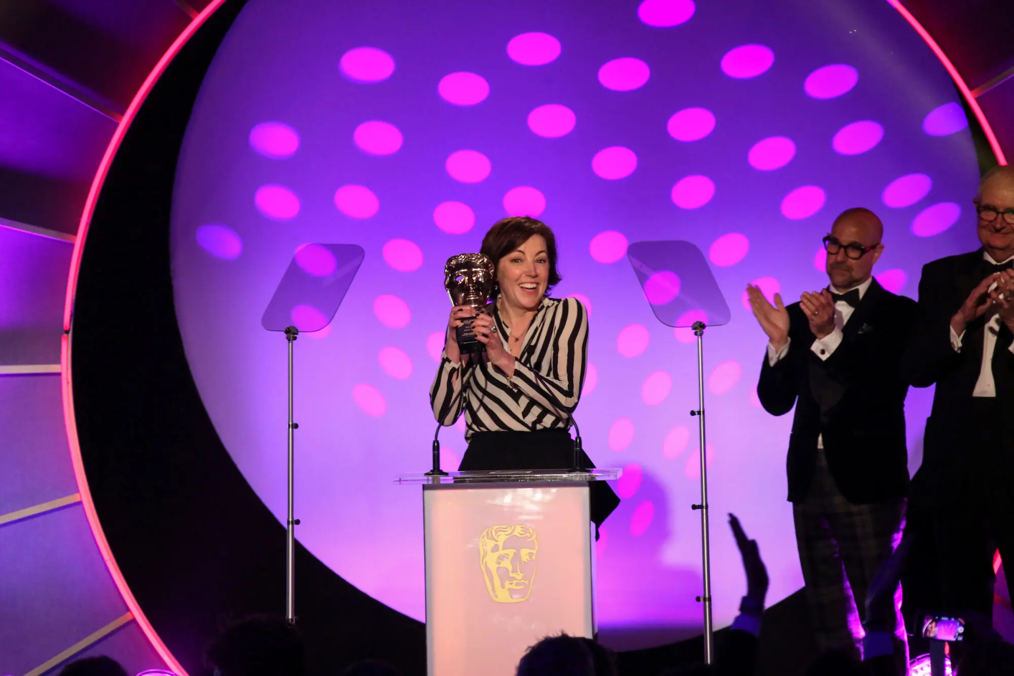A white woman with bobbed dark brown hair. She is wearing a black and white striped shirt and is holding a BAFTA award.