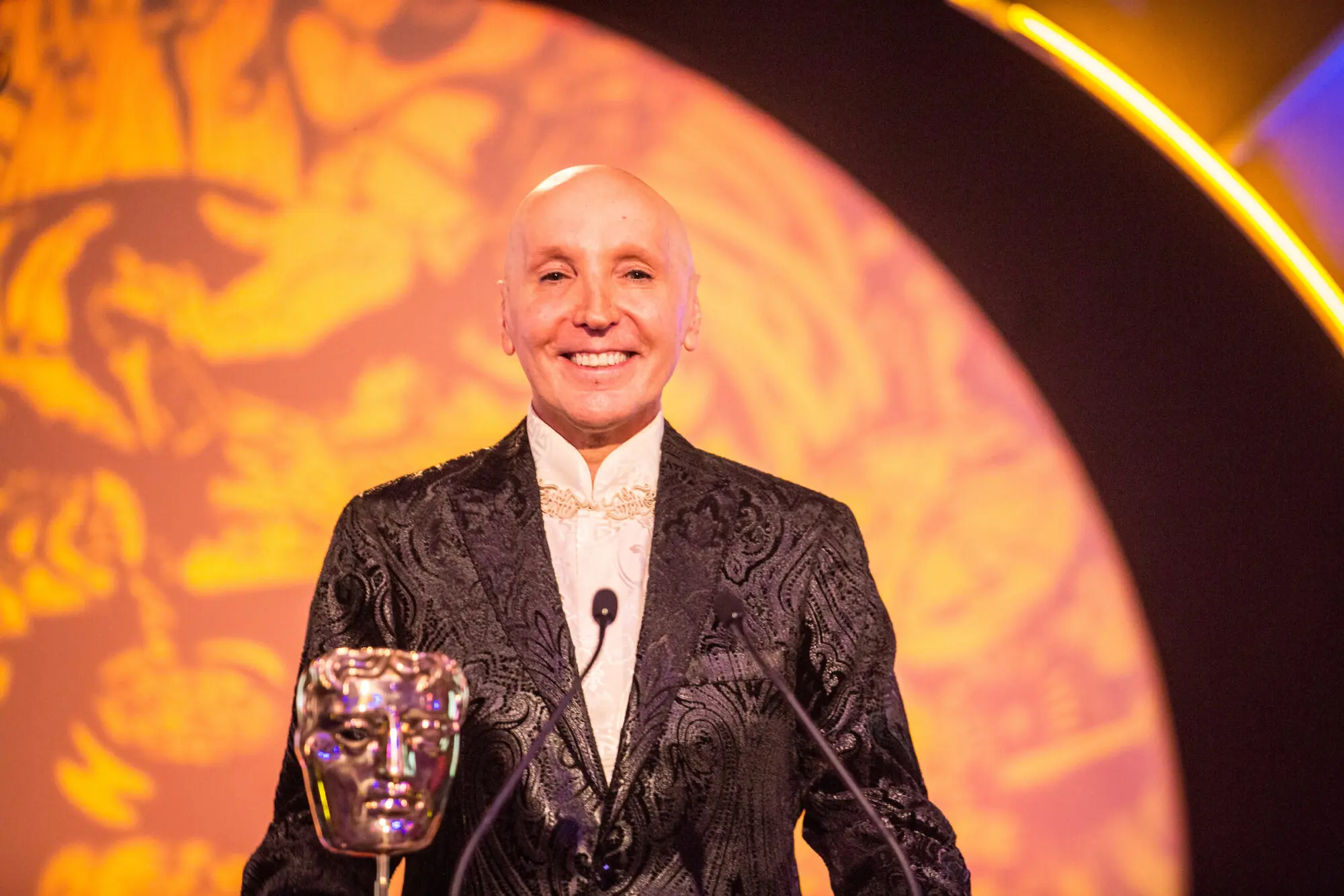 A bald white man wearing a dark suit embroidered with gold applique. He is smiling and standing with a BAFTA behind two microphones.
