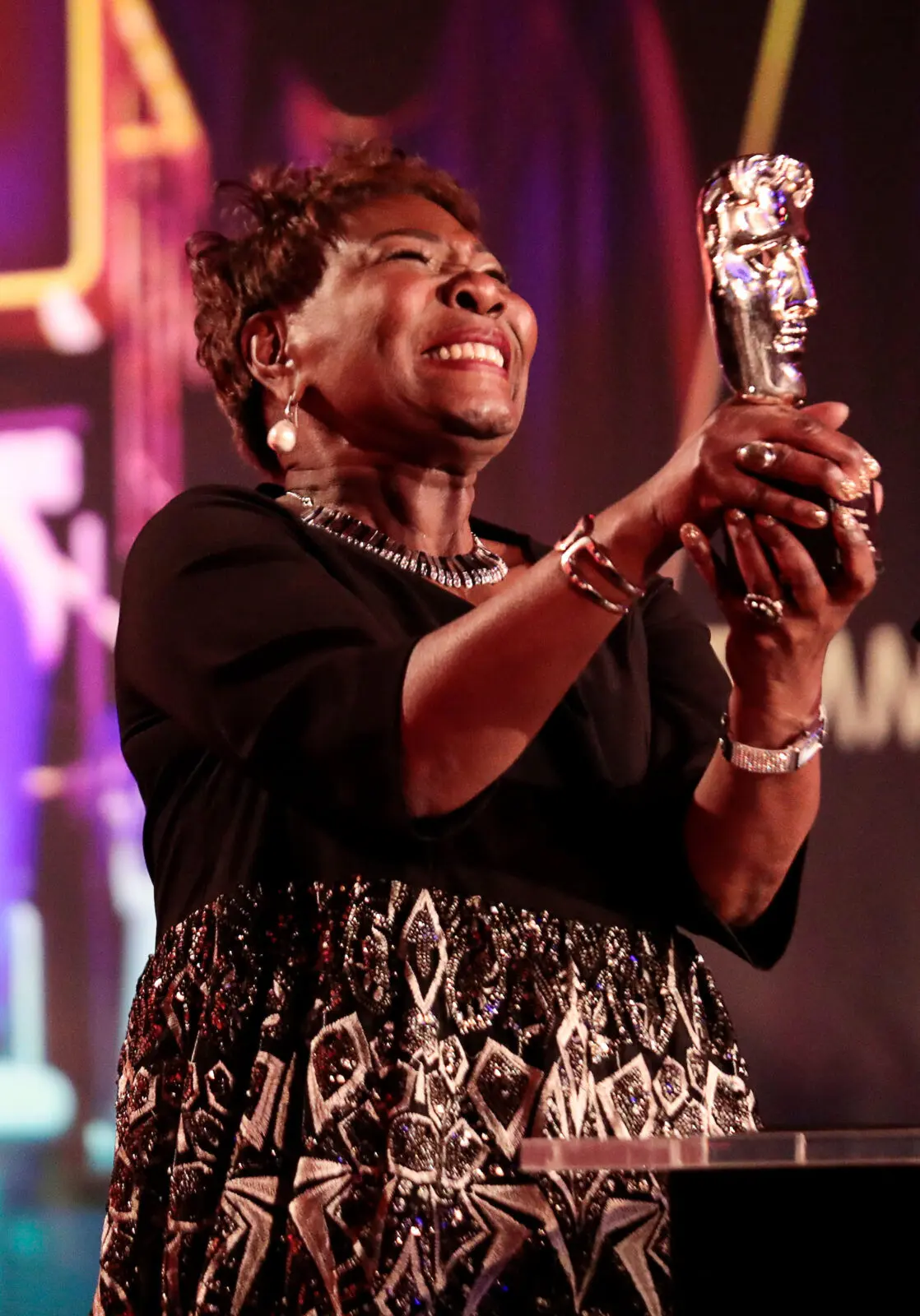 A black woman with a short pixie cut wearing a black top and gold patterned skirt. She is holding a BAFTA award.