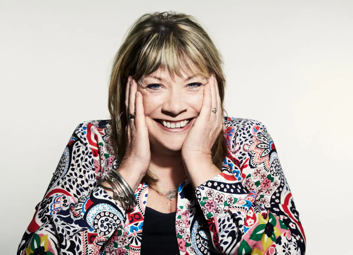 A white woman with a blonde bob and fringe. She is wearing a black top, patterned shirt, multiple silver bracelets, and is posing with her head in her hands.