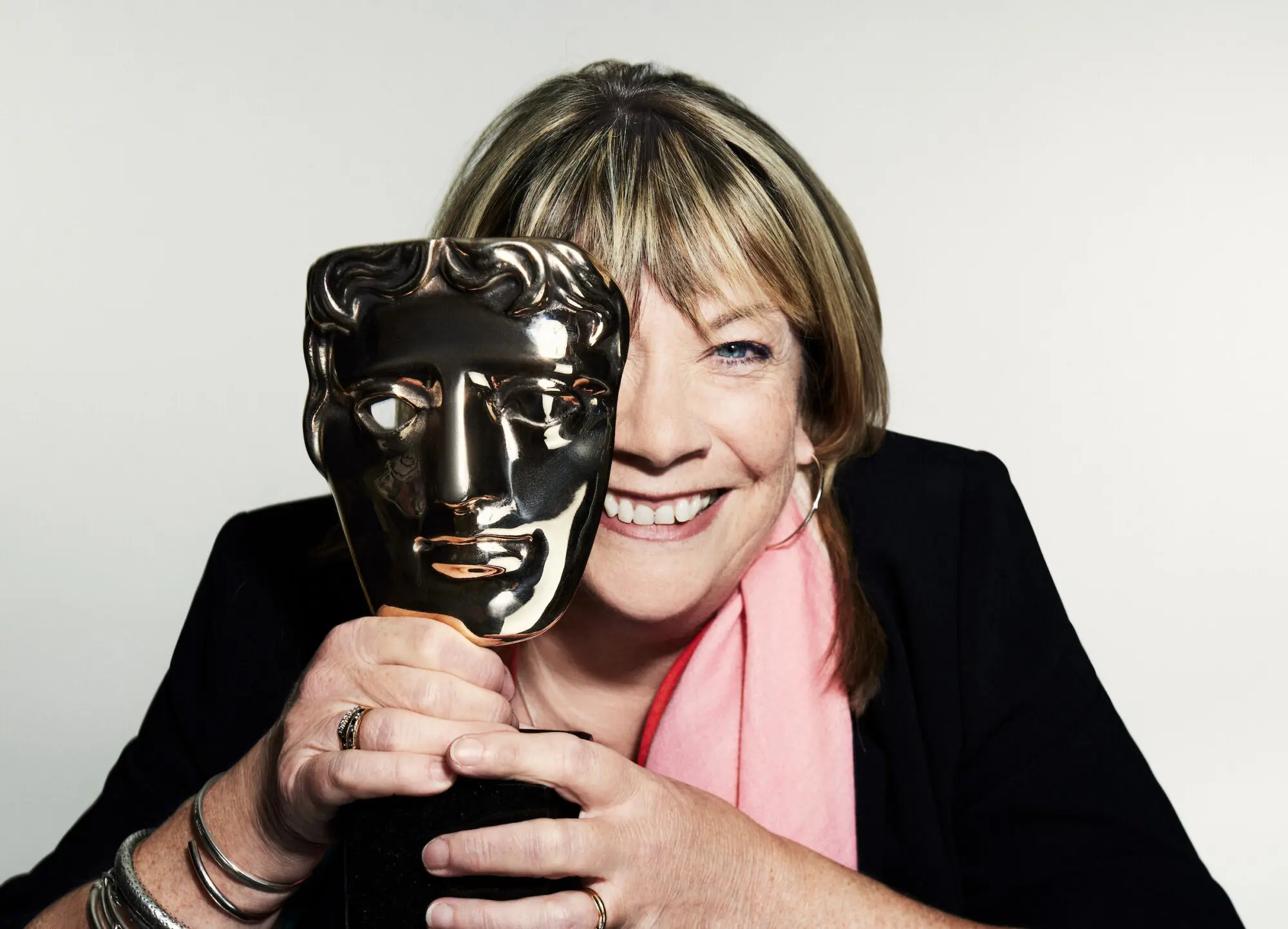 A white woman with a blonde bob and fringe. She is wearing a black jacket, pink scarf and is holding a BAFTA mask in front of her eye.